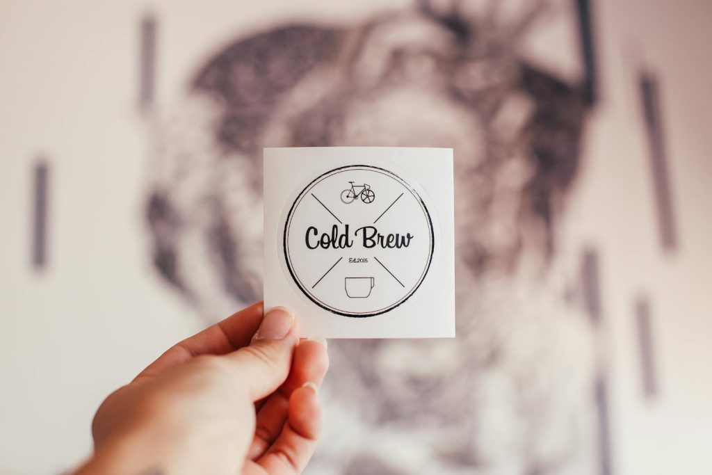 Unrecognizable person demonstrating coffee shop badge with Cold Brew inscription and creative design in hand while standing on blurred background